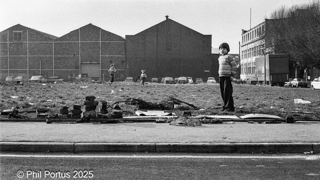 A young boy stands on a patch of debris near large industrial buildings and two other children in the background.
