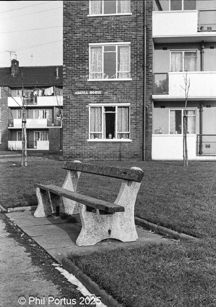 A black and white view shows a bench in front of a brick building with adjacent apartment blocks.