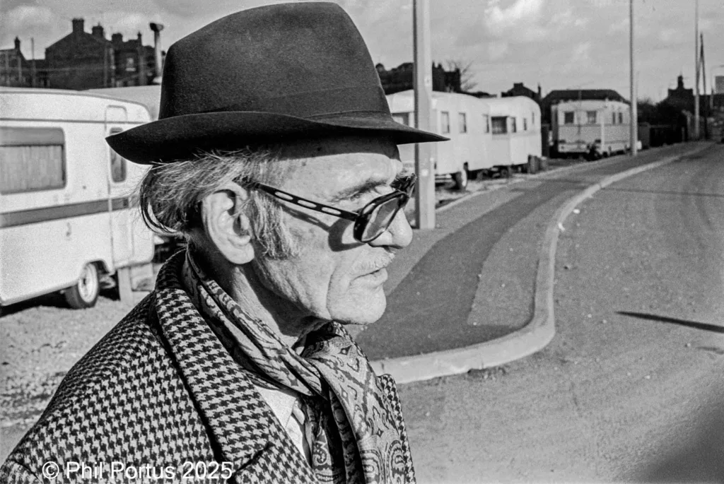 A man wearing a hat, glasses, and scarf looks off into the distance along a paved road.