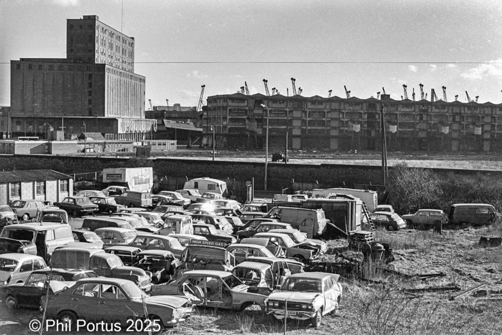 A black and white scene shows a large junkyard filled with numerous old cars and industrial buildings in the background.