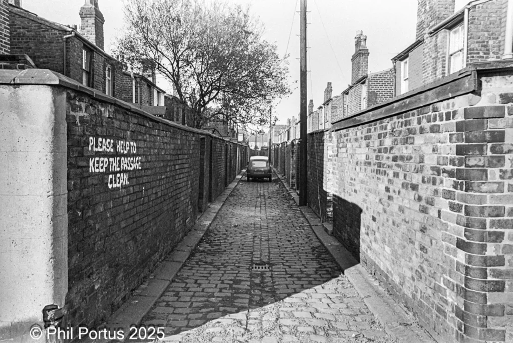 A narrow brick passage with a vehicle is flanked by tall brick walls and houses in black and white.