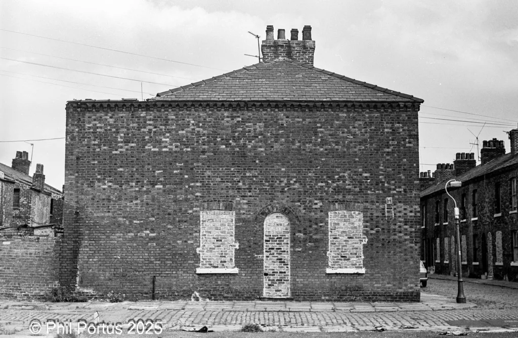 A black and white image shows a brick building with boarded windows and a steeply pitched roof.