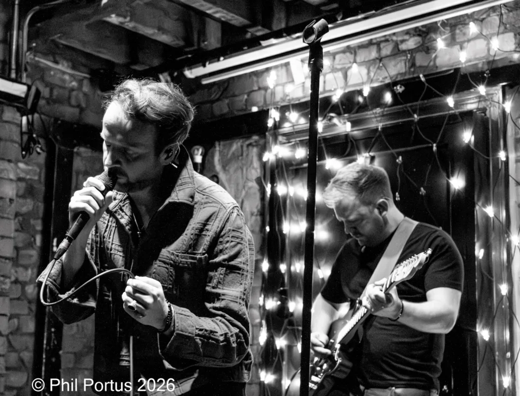 Two musicians perform on a stage with string lights behind them in black and white.