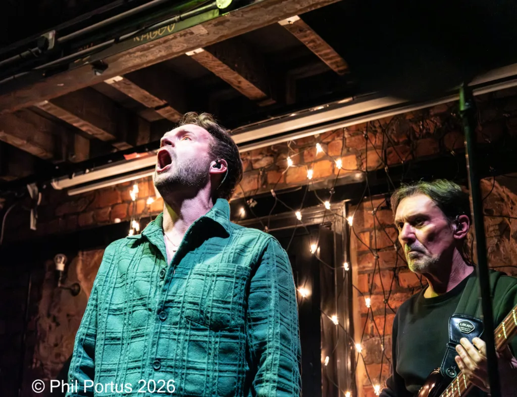 Two musicians perform on a dimly lit stage with exposed brick and string lights.