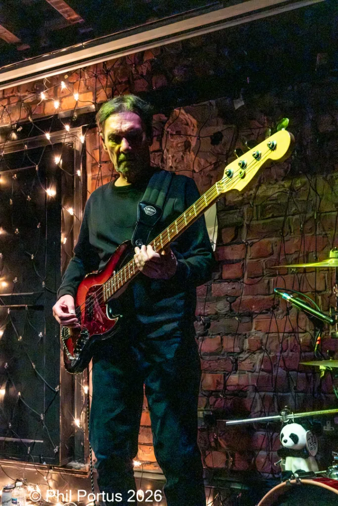 A musician plays a dark red electric bass guitar while standing in front of a brick wall with string lights.
