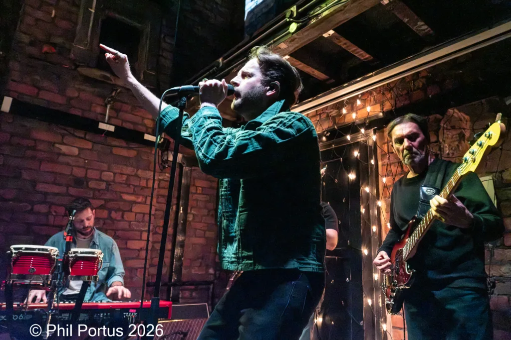 Three musicians perform on a stage with exposed brick walls and string lights.