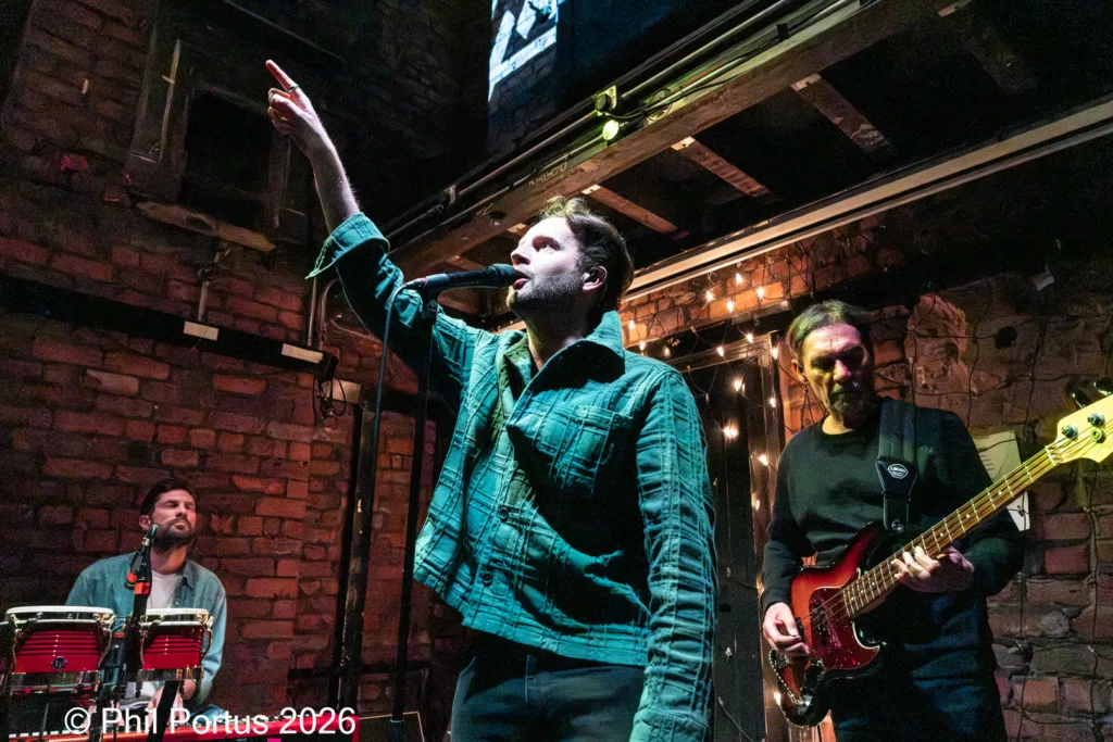 Three musicians perform on a brick stage under warm string lights.