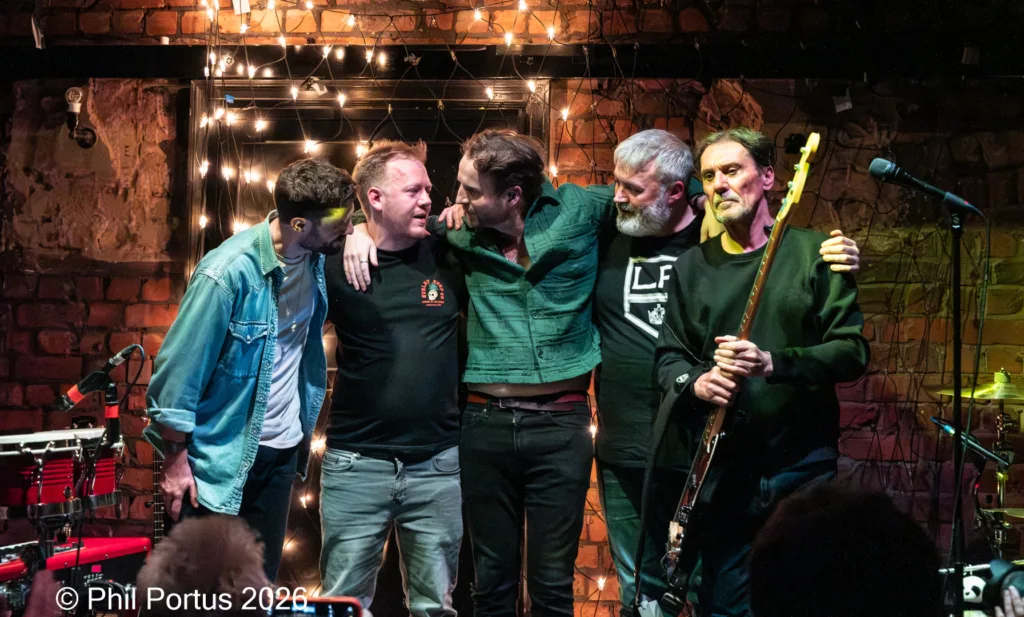 Five men stand close together against a brick wall decorated with string lights.